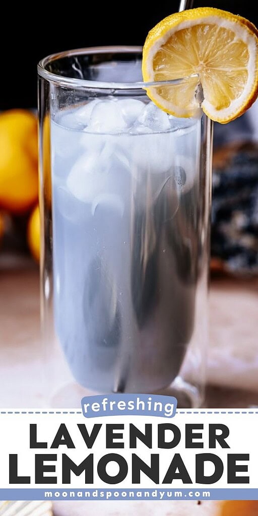 A glass of lavender lemonade with ice, garnished with a lemon slice, sits on a table with lemons and lavender in the background—perfect inspiration if you're searching for a refreshing lavender lemonade recipe.
