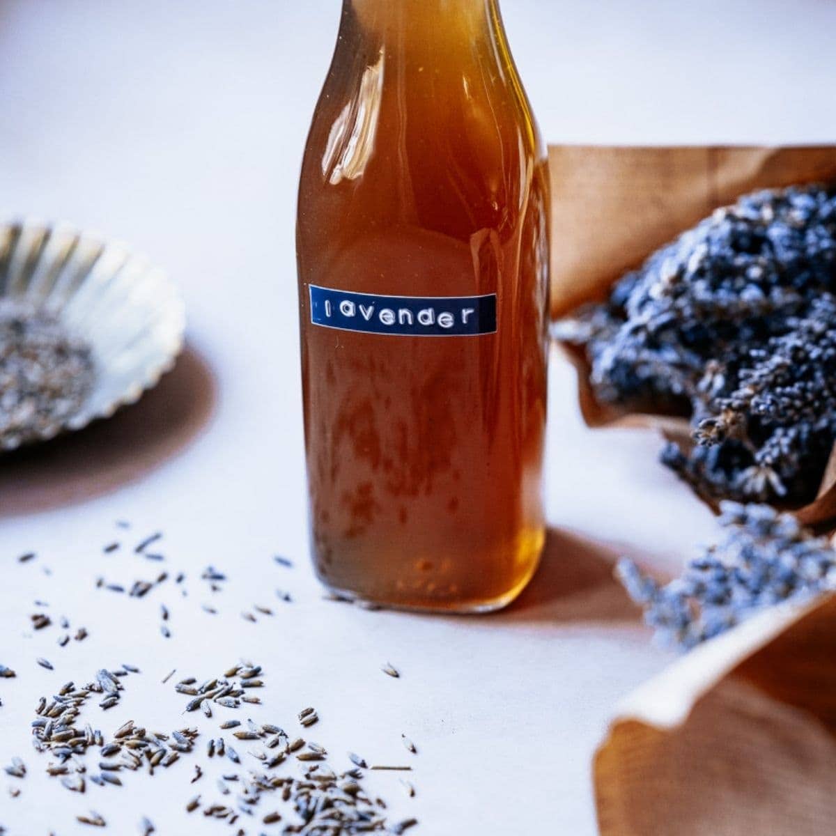 A bottle labeled "lavender" sits on a table among scattered lavender seeds and dried lavender bunches, hinting at the beginnings of a delightful lavender simple syrup recipe.