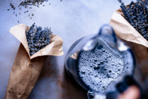 Two paper-wrapped bundles of dried lavender and a glass pitcher of soapy water are arranged on a light surface—perfect inspiration for your next lavender lemonade recipe.