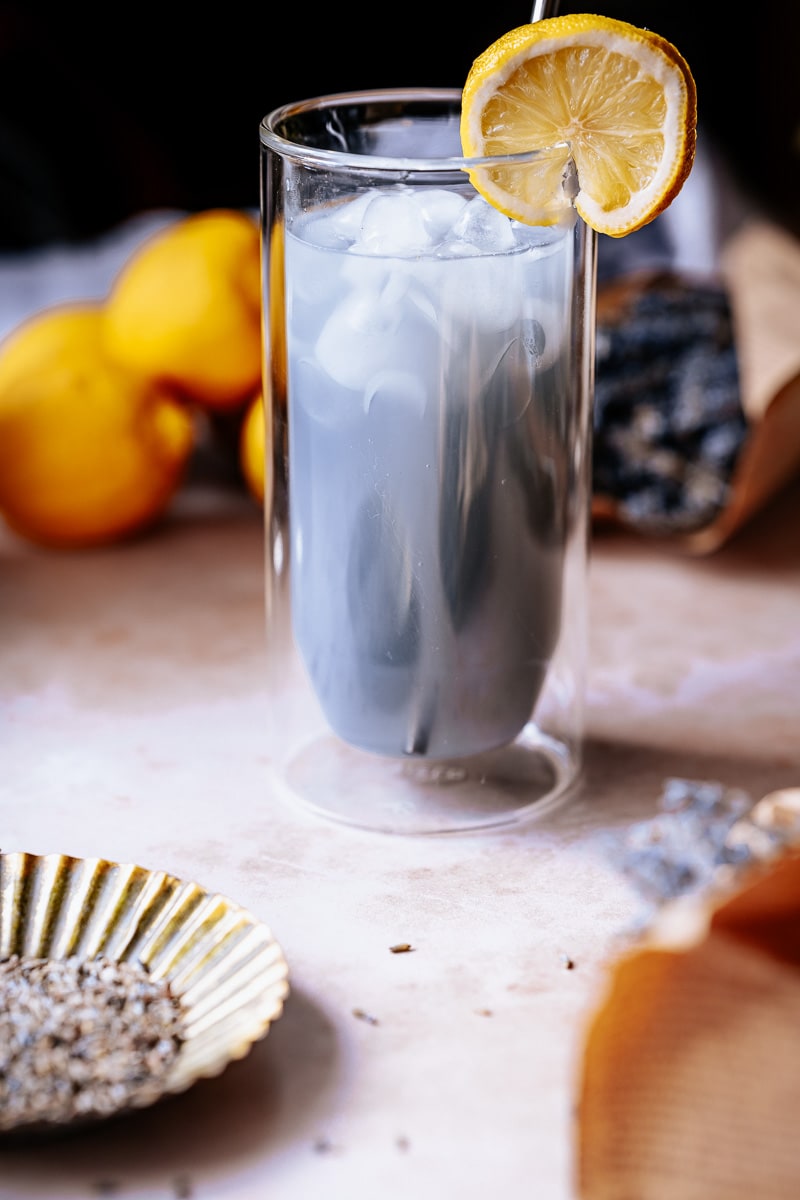 A lemon slice garnishes a tall clear glass filled with ice and purple lavender lemonade.