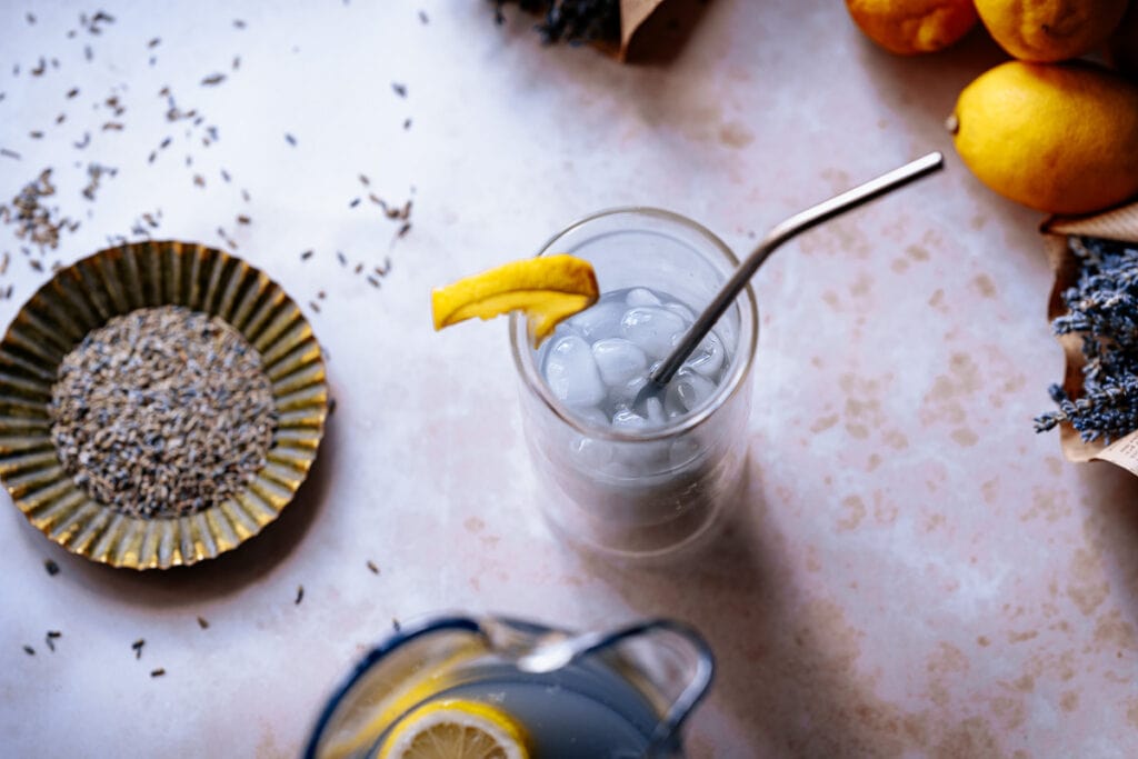 A glass of iced lavender lemonade with a lemon wedge and metal straw sits on a light surface, surrounded by fresh lemons, lavender, and a dish of dried lavender—perfect inspiration if you’re looking for a delicious lavender lemonade recipe.