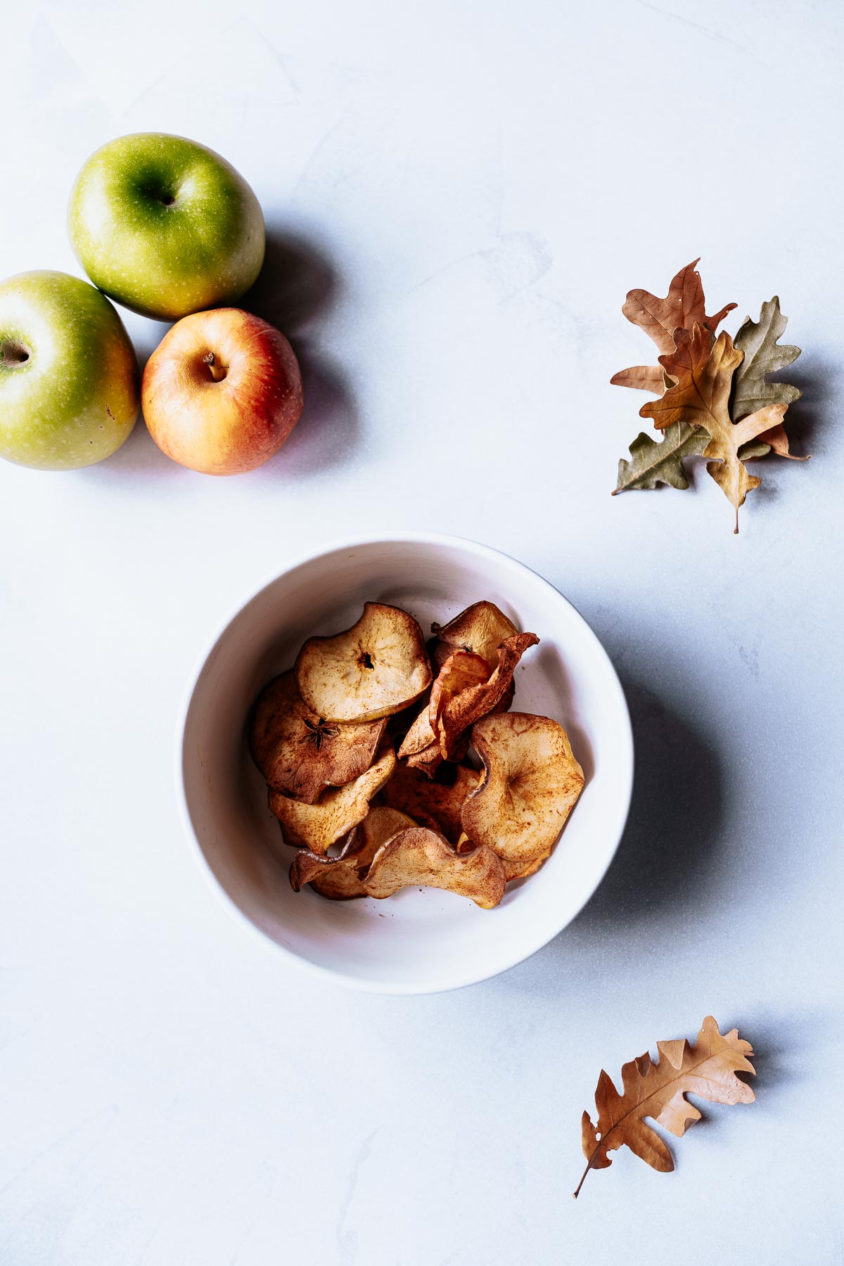 a small bowl of air fryer apple chips surrounded by green and red apples cinnamon sticks and fall leaves
