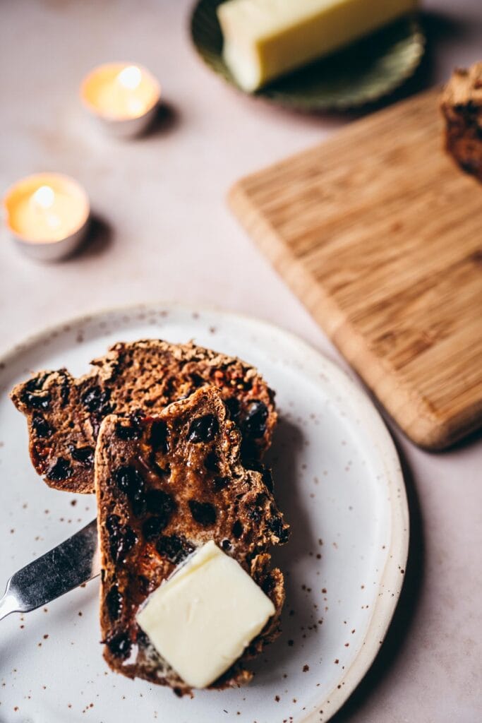 slices of warm irish gluten free barmbrack rest on a speckled ceramic plate alongside pats of butter