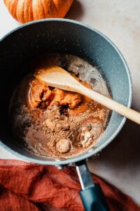 A saucepan with pumpkin puree, spices, and a wooden spoon sits on the countertop, ready for making homemade pumpkin spice syrup. Nearby, a pumpkin and a red cloth add to the cozy autumn atmosphere—perfect for creating your own pumpkin spice coffee syrup recipe at home.