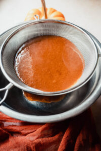 A fine mesh strainer sits over a metal bowl, filtering a brown liquid of homemade pumpkin spice syrup. A small pumpkin and an orange cloth are visible in the background.