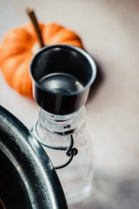 A close-up of a glass bottle filled with homemade pumpkin spice syrup with a metal cap next to a small orange pumpkin on a light surface.