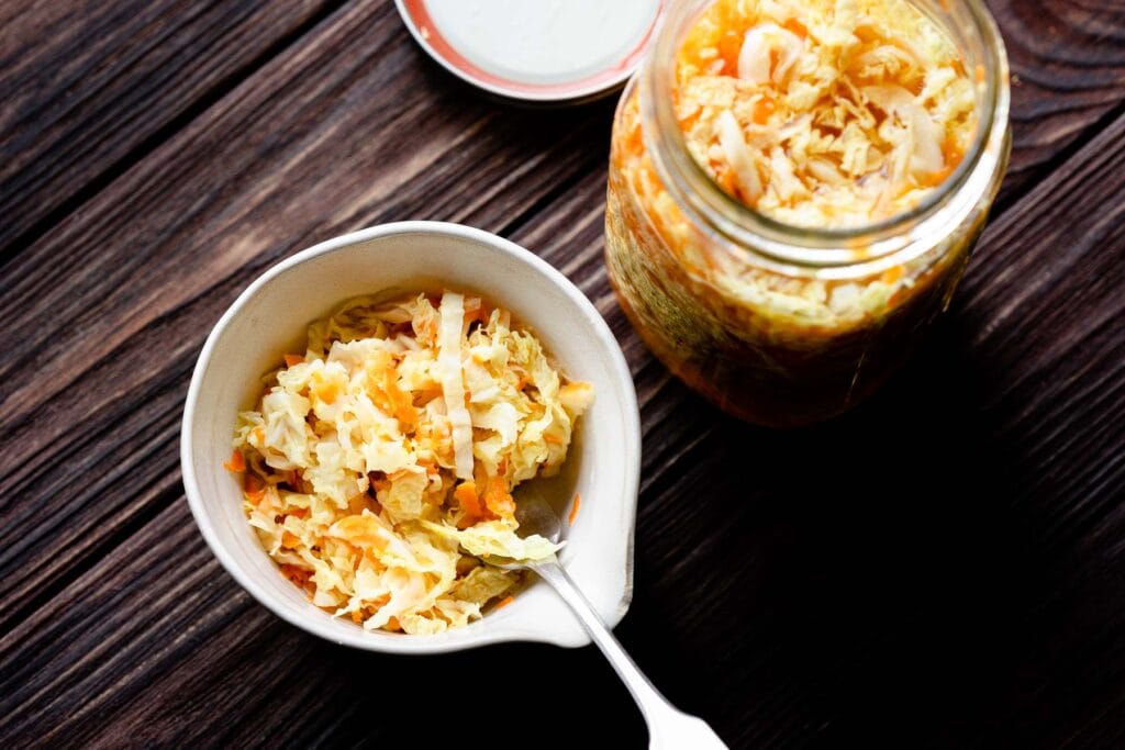 A bowl and a jar of sauerkraut with shredded pickled cabbage and carrots sit on a dark wooden surface, with a spoon in the bowl.