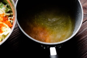 A pot of simmering broth sits on a dark wooden surface next to a bowl of chopped vegetables, including carrots and tangy pickled cabbage.