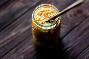 A glass jar filled with homemade pickled cabbage ferment, possibly sauerkraut, sits on a dark wooden surface with a wooden utensil inside.