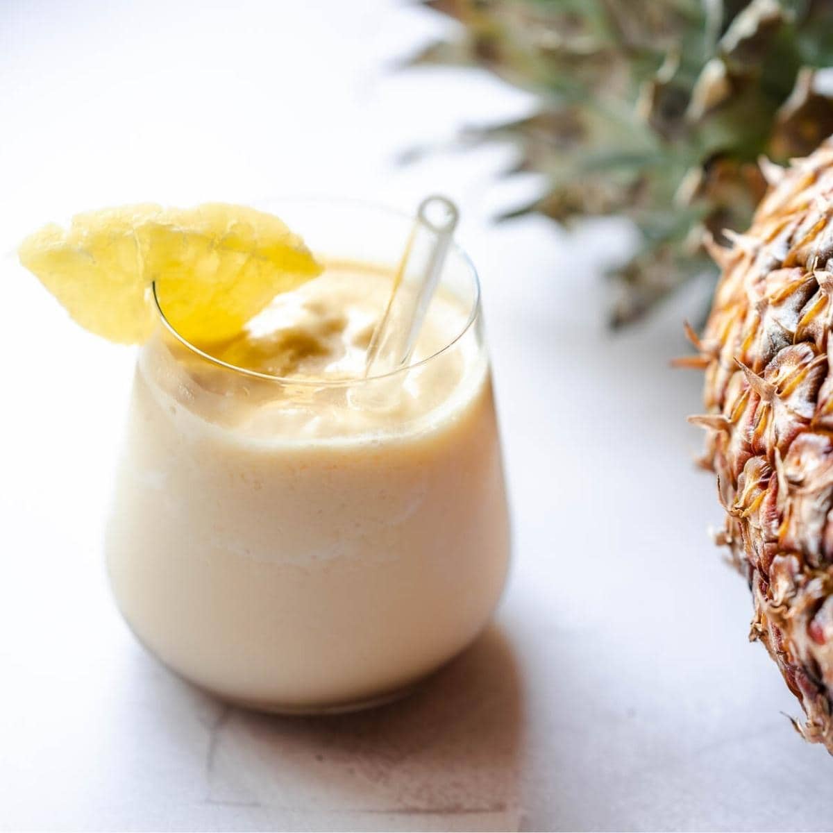 a clear glass filled with pale yellow frozen pineapple smoothie garnished with dried pineapple ring and a glass straw