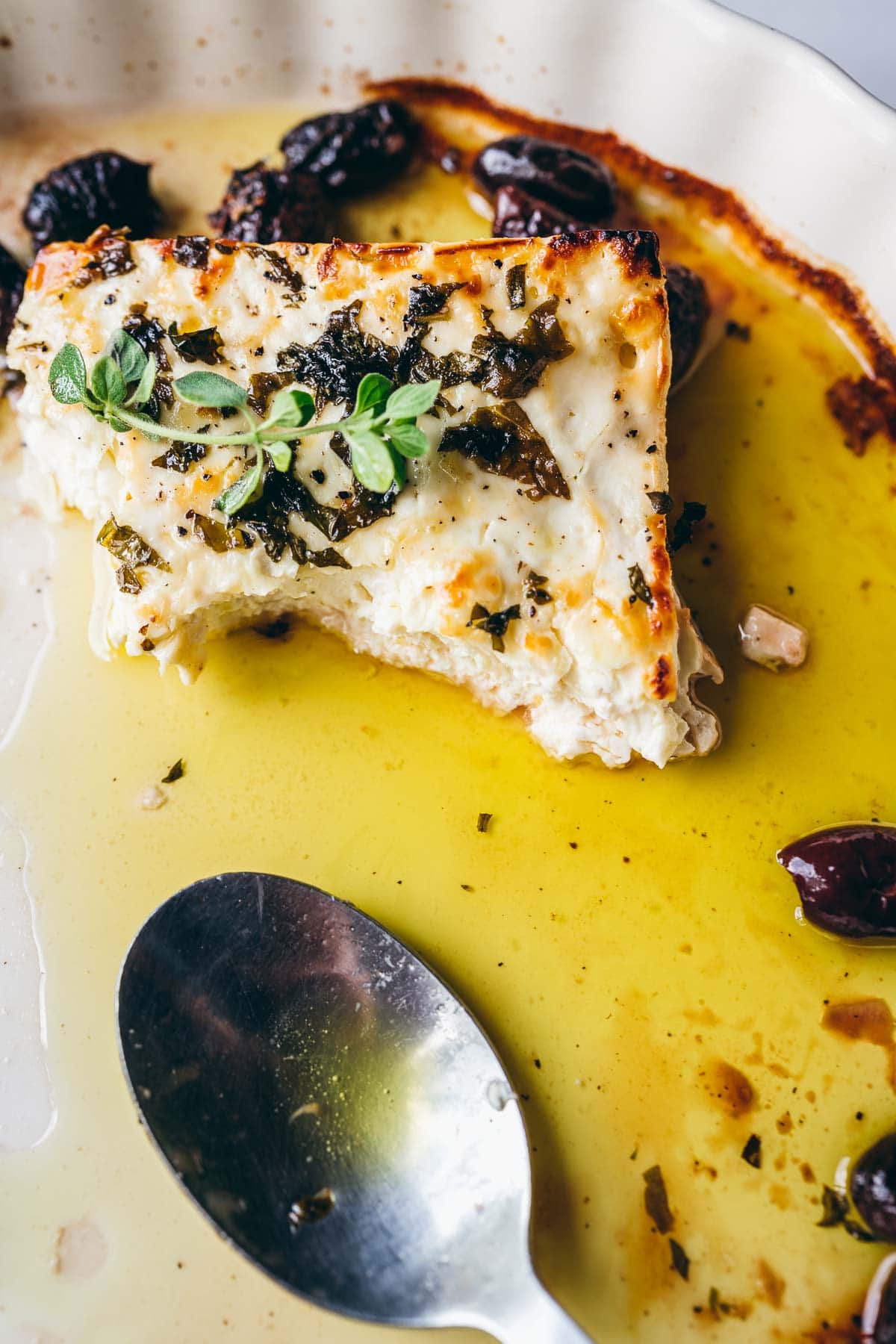 a spoon rests in a baking dish with olive oil wilted olives and freshly baked feta cheese that was just cut open