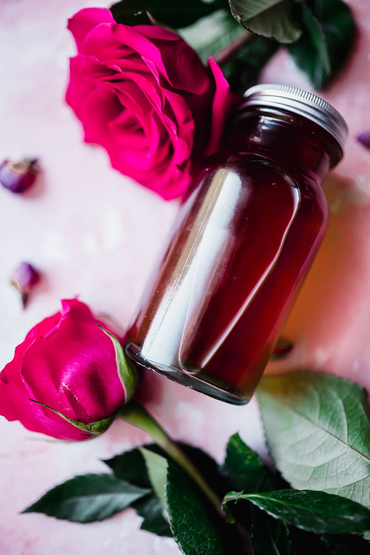 a bottle of rose water made at home is surrounding by two bright pink rose blossoms.