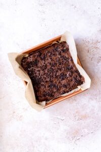 A square baking pan lined with parchment paper holds a batch of decadent black bean brownies topped with chocolate chunks.