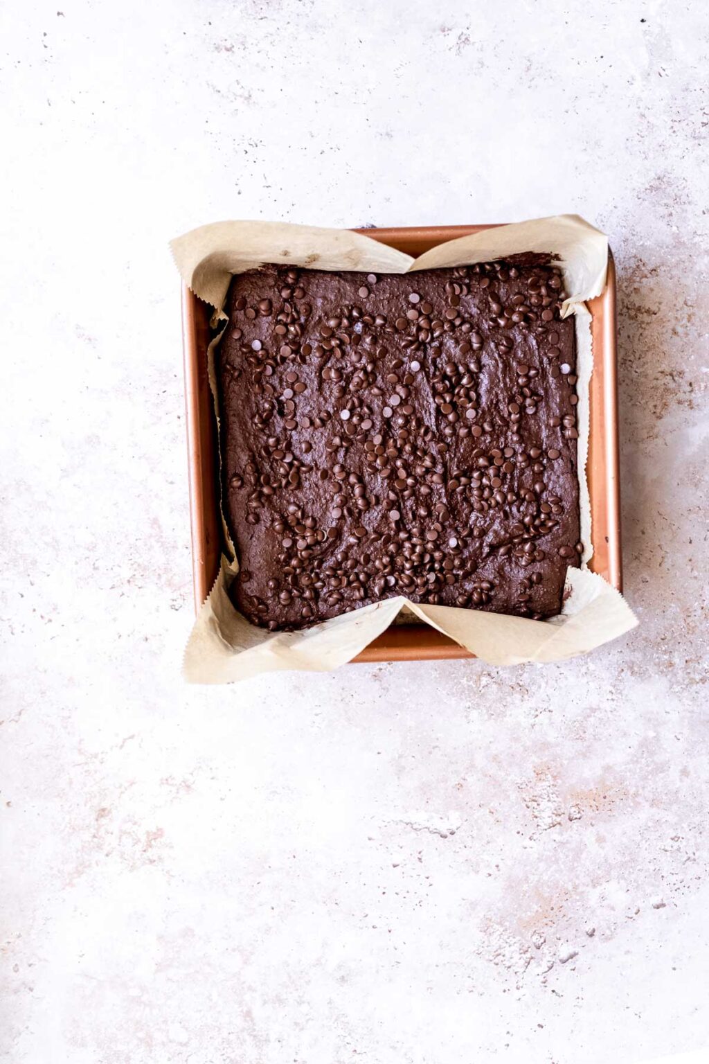 A square baking pan lined with parchment paper holds unbaked black bean brownie batter topped with chocolate chips, placed on a light-colored surface.