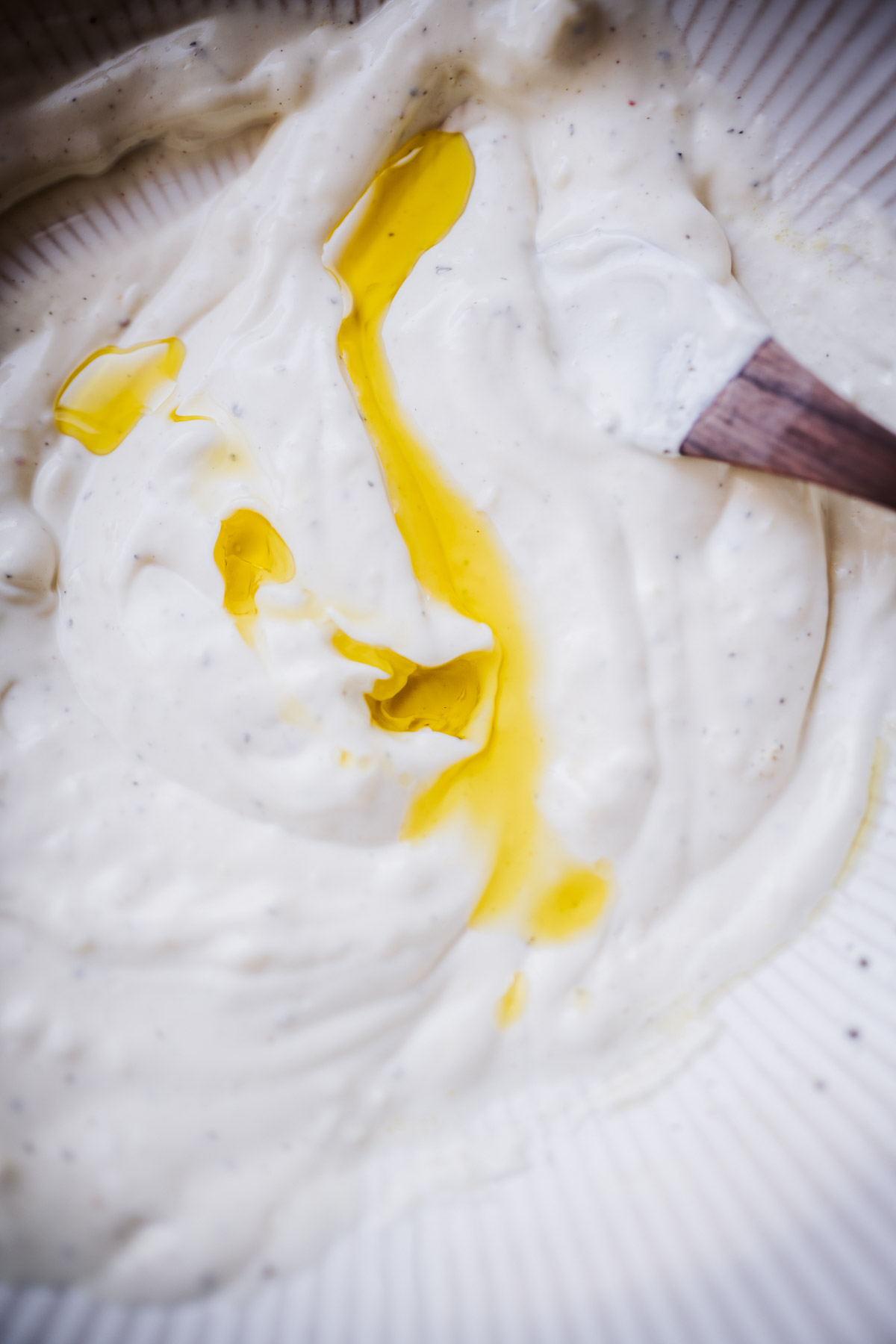 a close view of a wooden spoon mixing aioli sauce drizzled with olive oil.