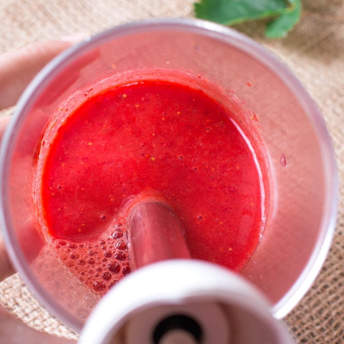 a bright red fruit puree in a glass cup with an immersion blender stuck in.