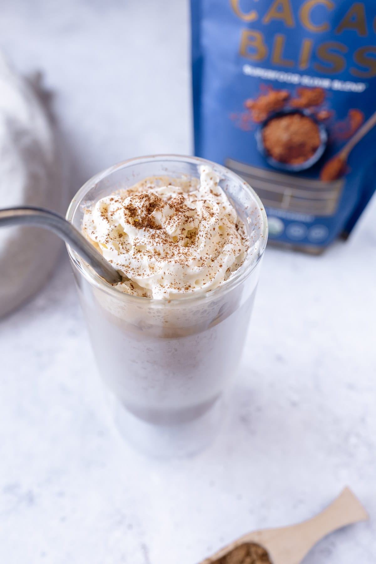 a side view of a tall clear glass filled with peanut butter cup latte topped with coconut whipped cream and dusting of earth echo cacao bliss powder.