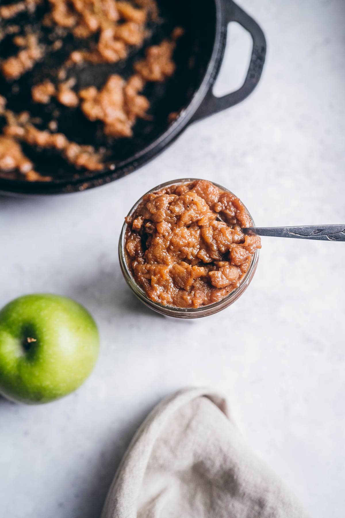 a jar of apple curd rests next to a green apple, cast iron pan and a tan napkin.