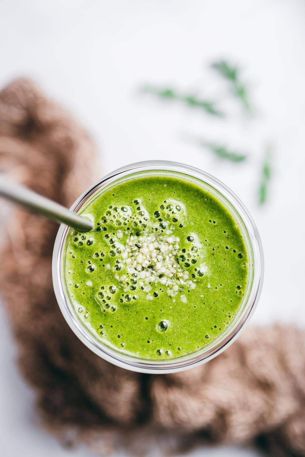 a bright green arugula smoothie recipe in a clear glass with a metal straw garnished with small white seeds.