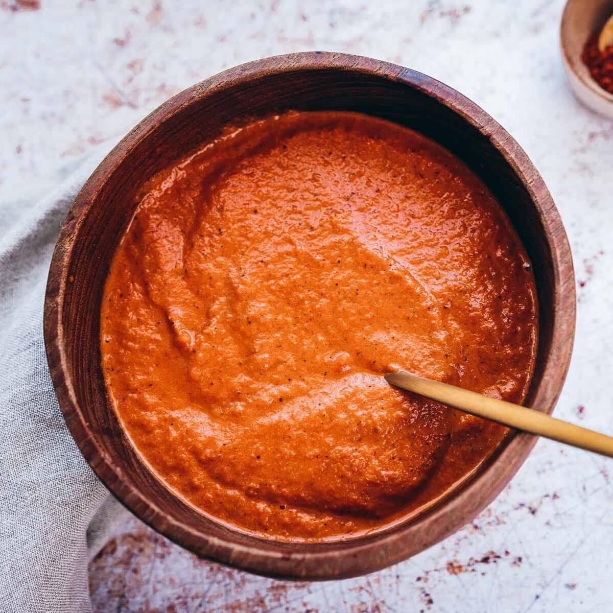 a teak wooden bowl filled with bright red salsa bravas sauce alongside a gold spoon.