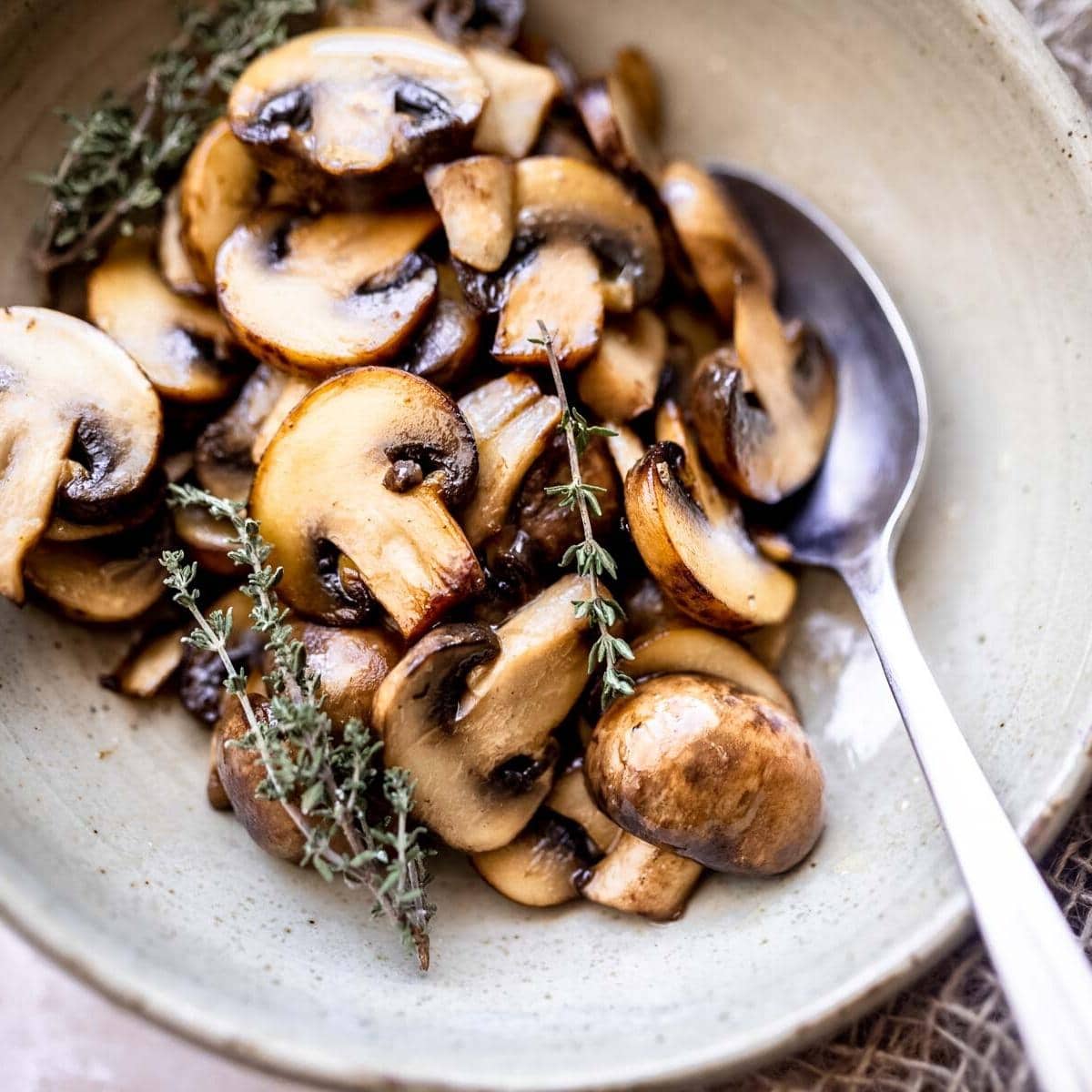 close shot of sauteed baby bella mushrooms in a ceramic bowl topped with a sprig of thyme.