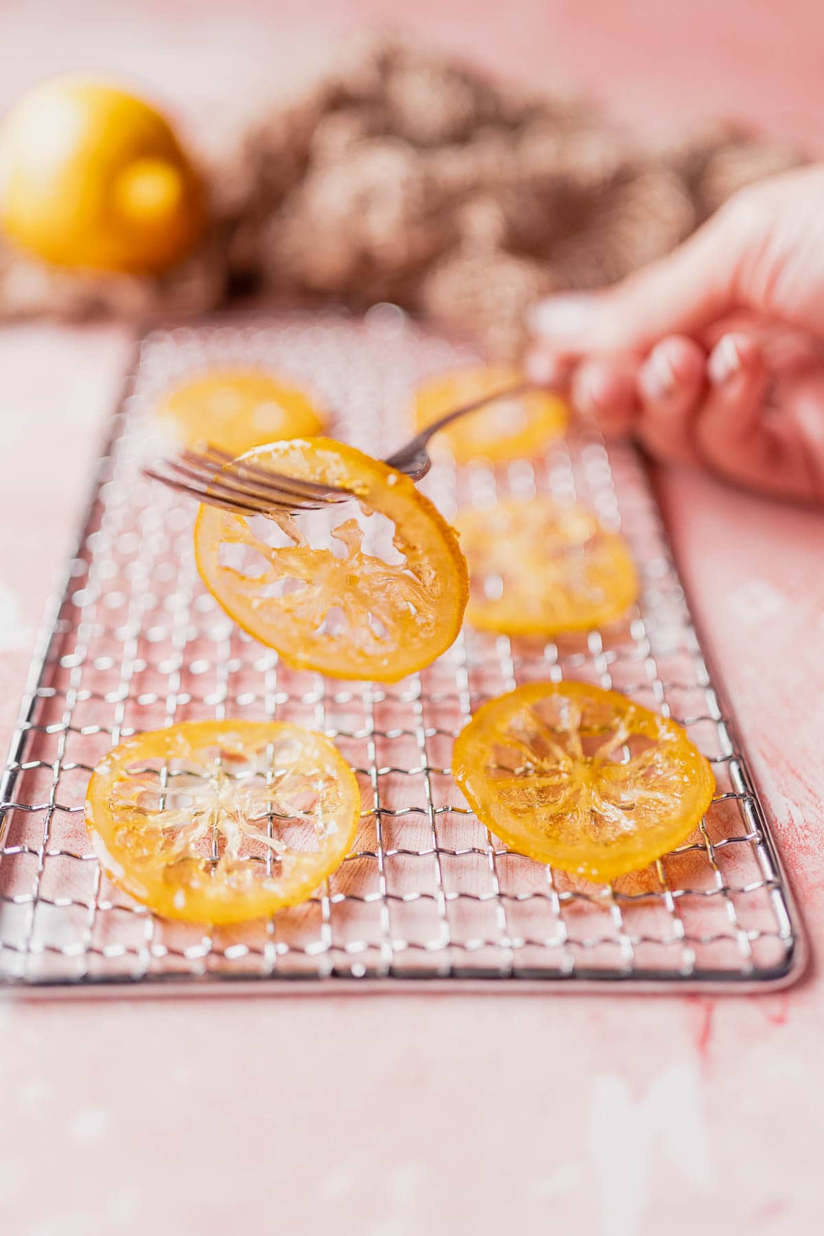 A hand rests on a pink table as it lifts a silver fork poked through a thin candied lemon slice.