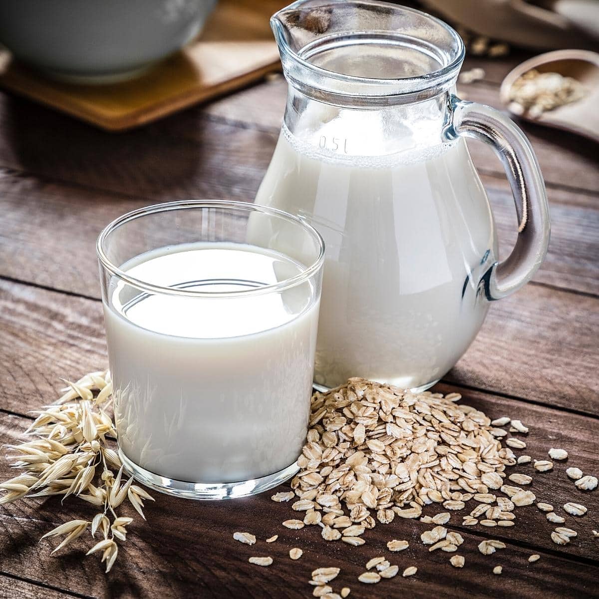 One clear jug and one clear glass filled with white oat milk resting on dark wooden table.