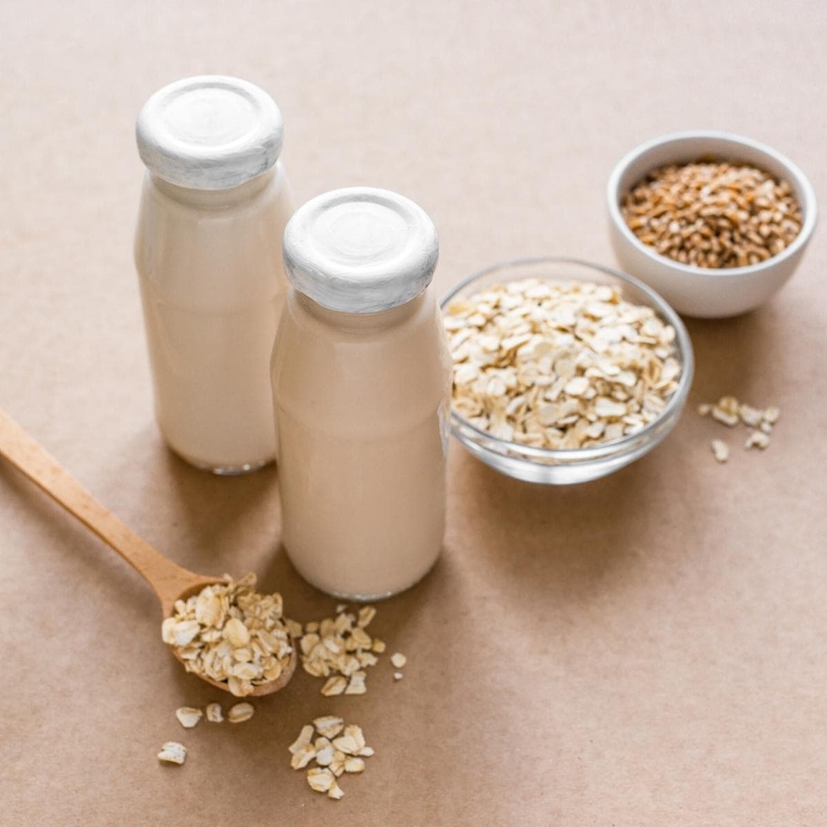 A pink table scattered with rolled oats and glasses of white oat milk with white lids.