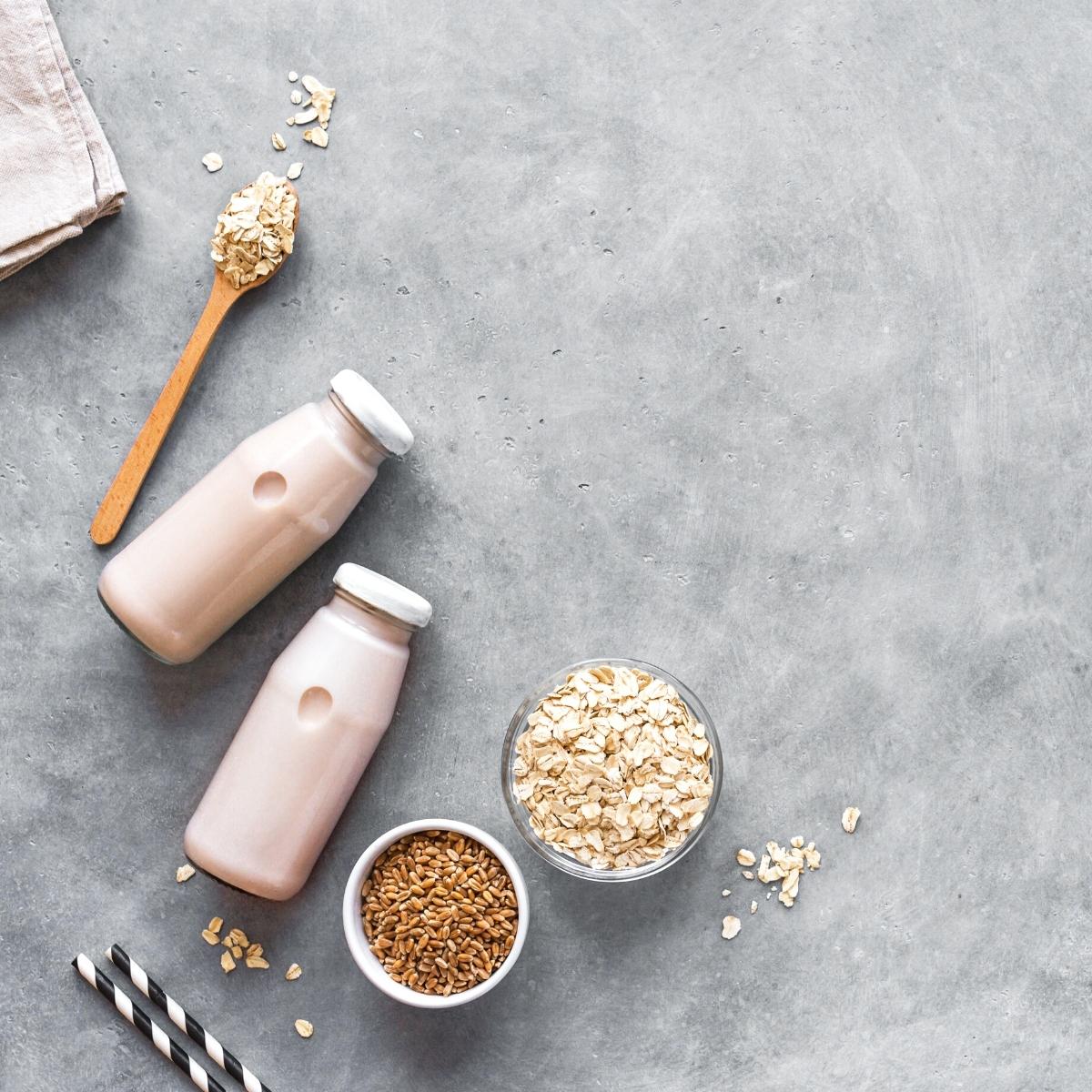 A gray backdrop sprinkled with rolled oats and bowls of steel cut oats, glasses of oat milk and black and white straws.