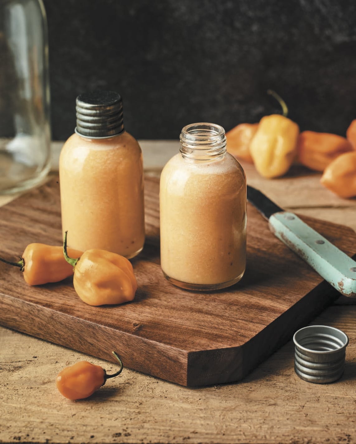 Glass bottles filled with orange sauce rest on a wood cutting board next to a knife and habanero chiles.