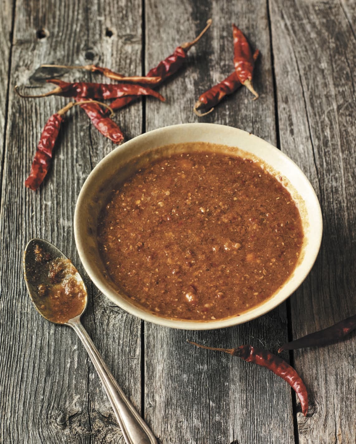 A small dish filled with homemade hot sauce resting on a wood table scattered with dried red chiles.