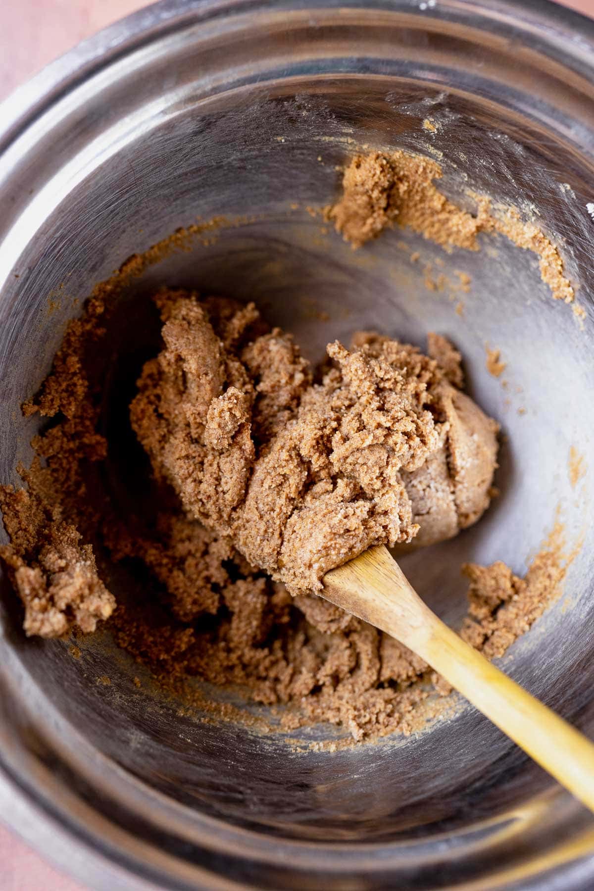 A wooden spoon stirs a brown dough in a silver mixing bowl.