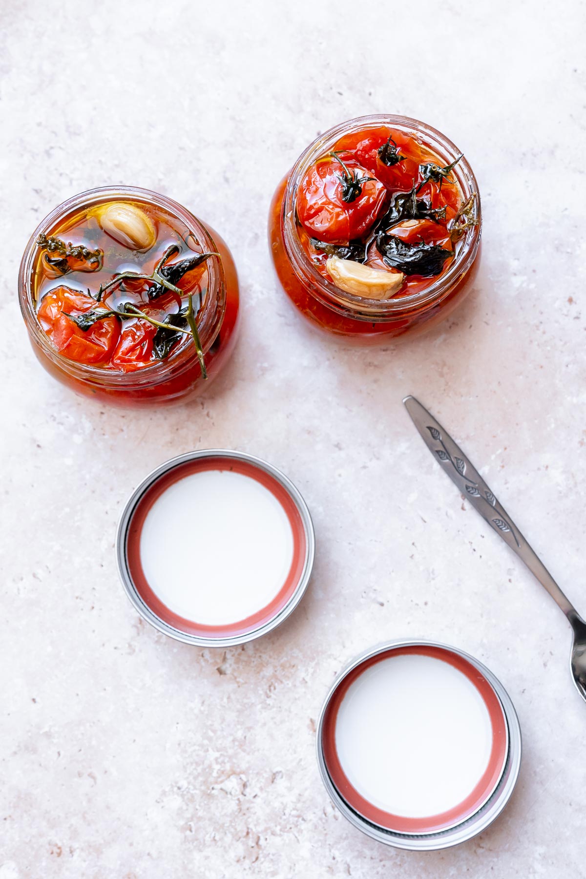 Two open clear jars filled with oil, herbs and cherry tomatoes.