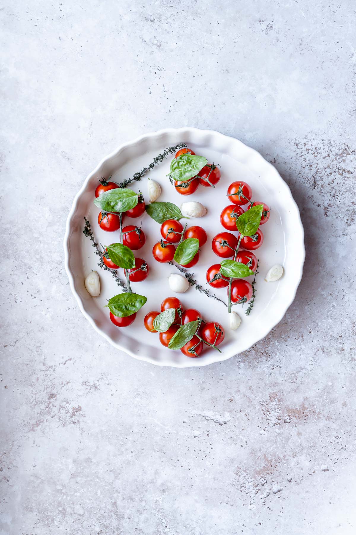 A round white baking dish filled with cherry tomatoes on the vine, fresh green herbs, and garlic cloves.