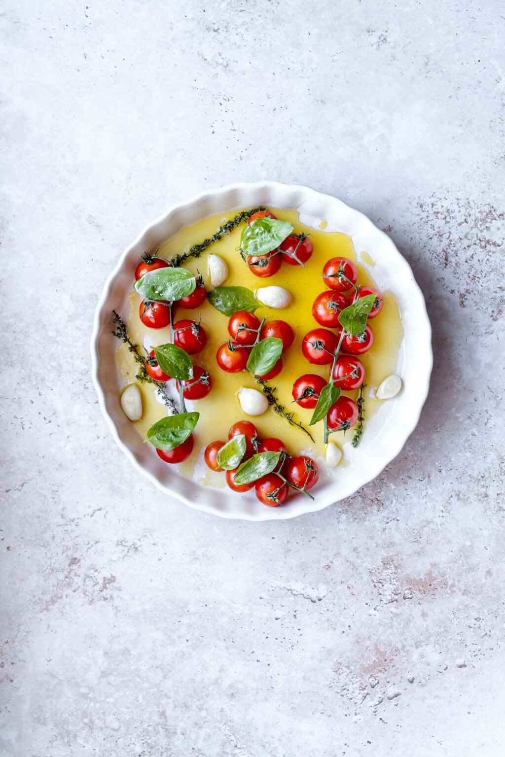 White dish with cherry tomatoes, basil leaves, garlic cloves, and sprigs of thyme in olive oil on a light surface, creating a delightful tomato confit.