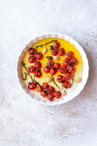 A round dish featuring a delightful tomato confit, with roasted cherry tomatoes on the vine, complemented by aromatic garlic cloves and fresh herbs gently infused in olive oil.