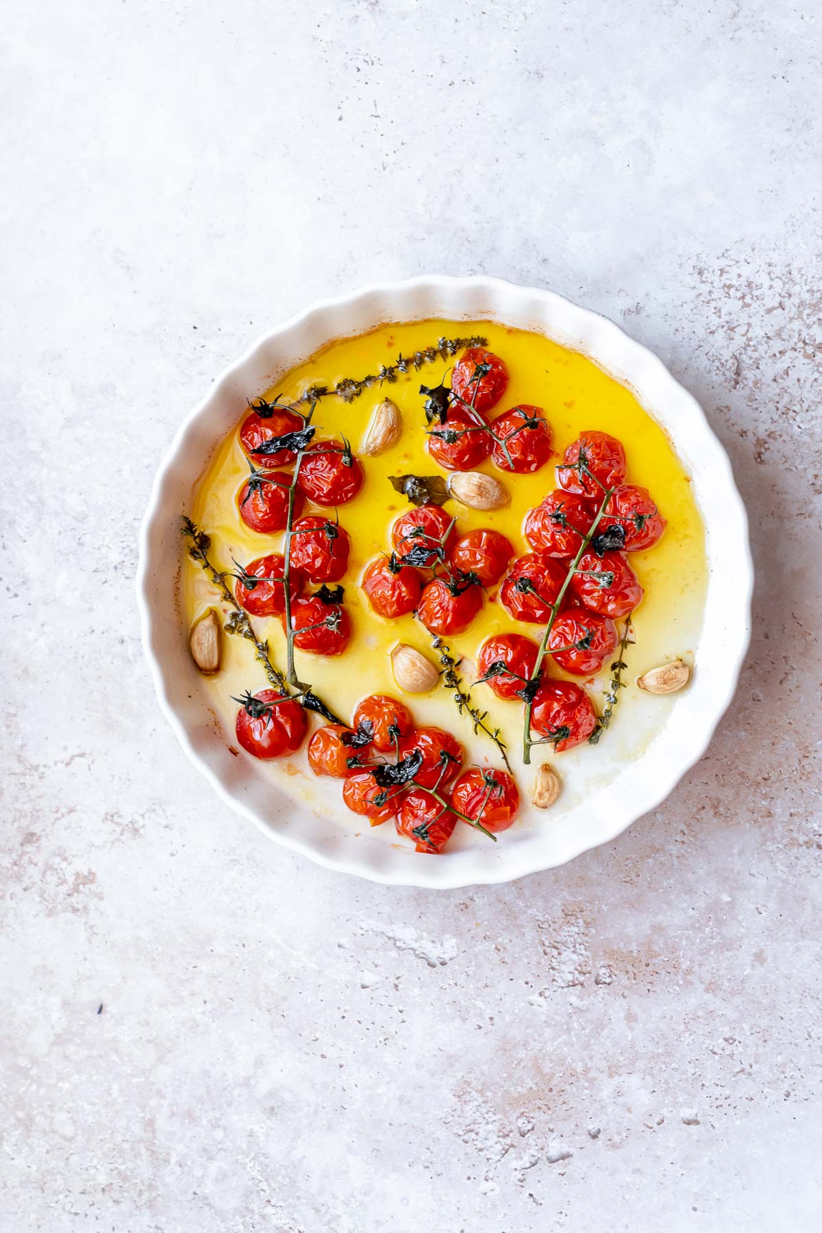 A round white baking dish filled with softened cherry tomatoes on the vine, golden garlic cloves and green herbs.