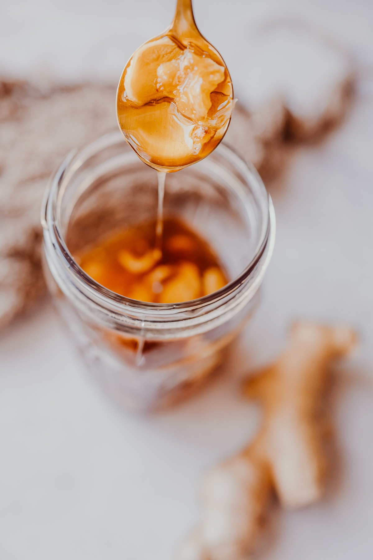 A golden spoon drizzling ginger root and honey into a clear jar.