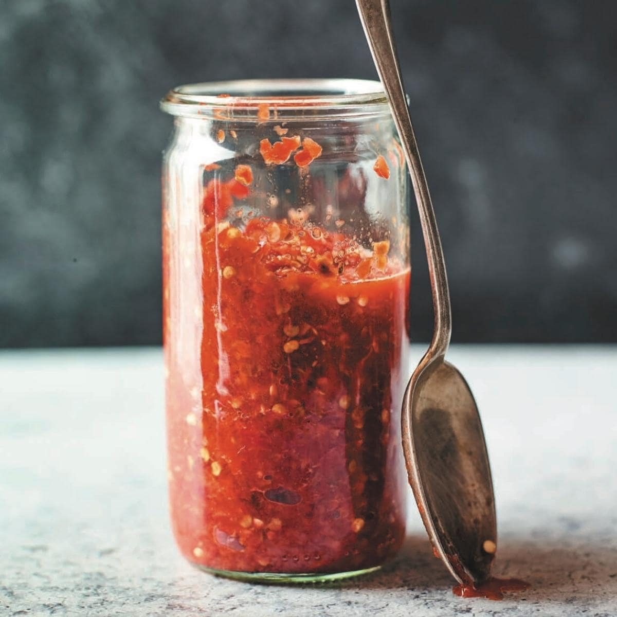 A clear jar filled with a red chile paste with a silver spoon resting against the jar.