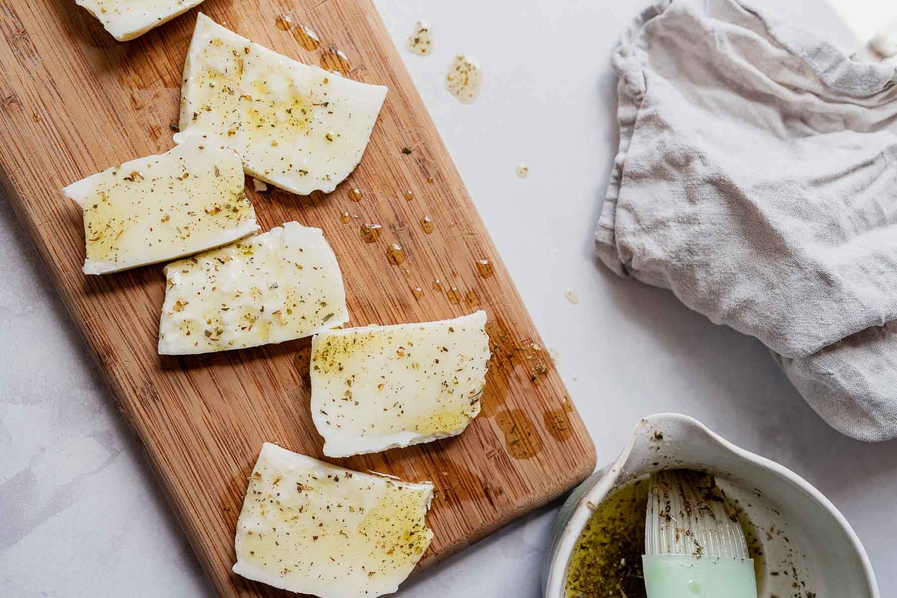 Slices of white cheese drizzled with oil rest on a small wooden cutting board.