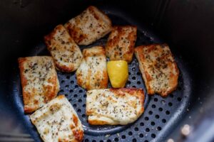 Pieces of cooked fish seasoned with herbs, complemented by crispy air fryer halloumi and a wedge of lemon, are arranged in an air fryer basket.