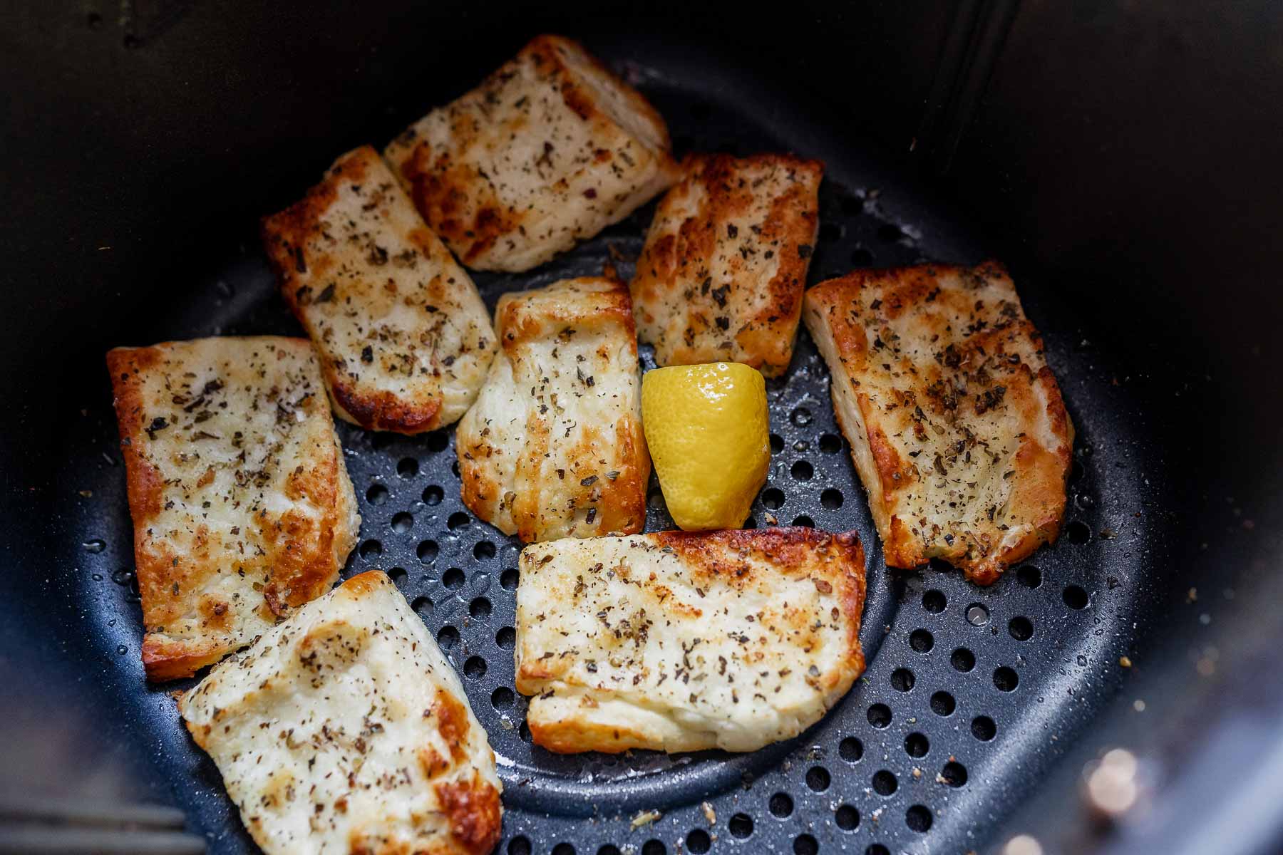 Slices of golden cheese rest in the bottom of an air fryer basket next to a fresh lemon wedge.