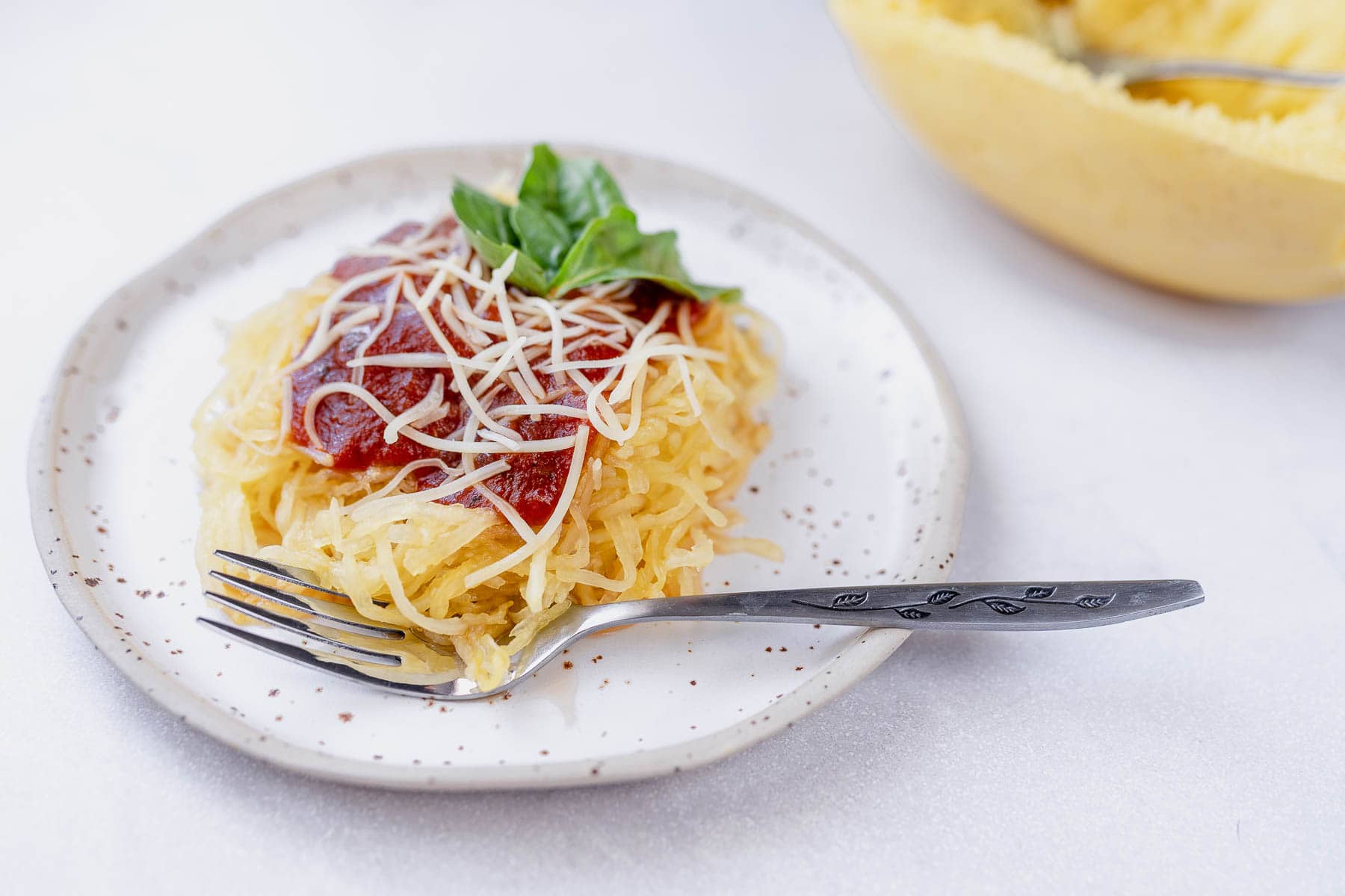 Spaghetti squash on a stoneware plate with a fork and marinara, parmesan and fresh basil on top, next to spaghetti squash with a fork.