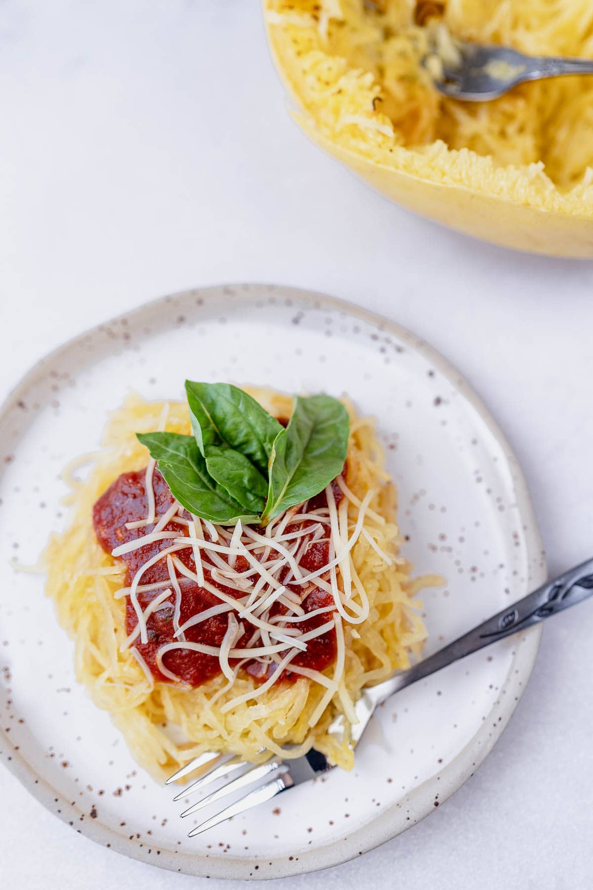 Overhead shot of squash on a stoneware plate and fork with marinara, parmesan and fresh basil on top, next to spaghetti squash with a fork.