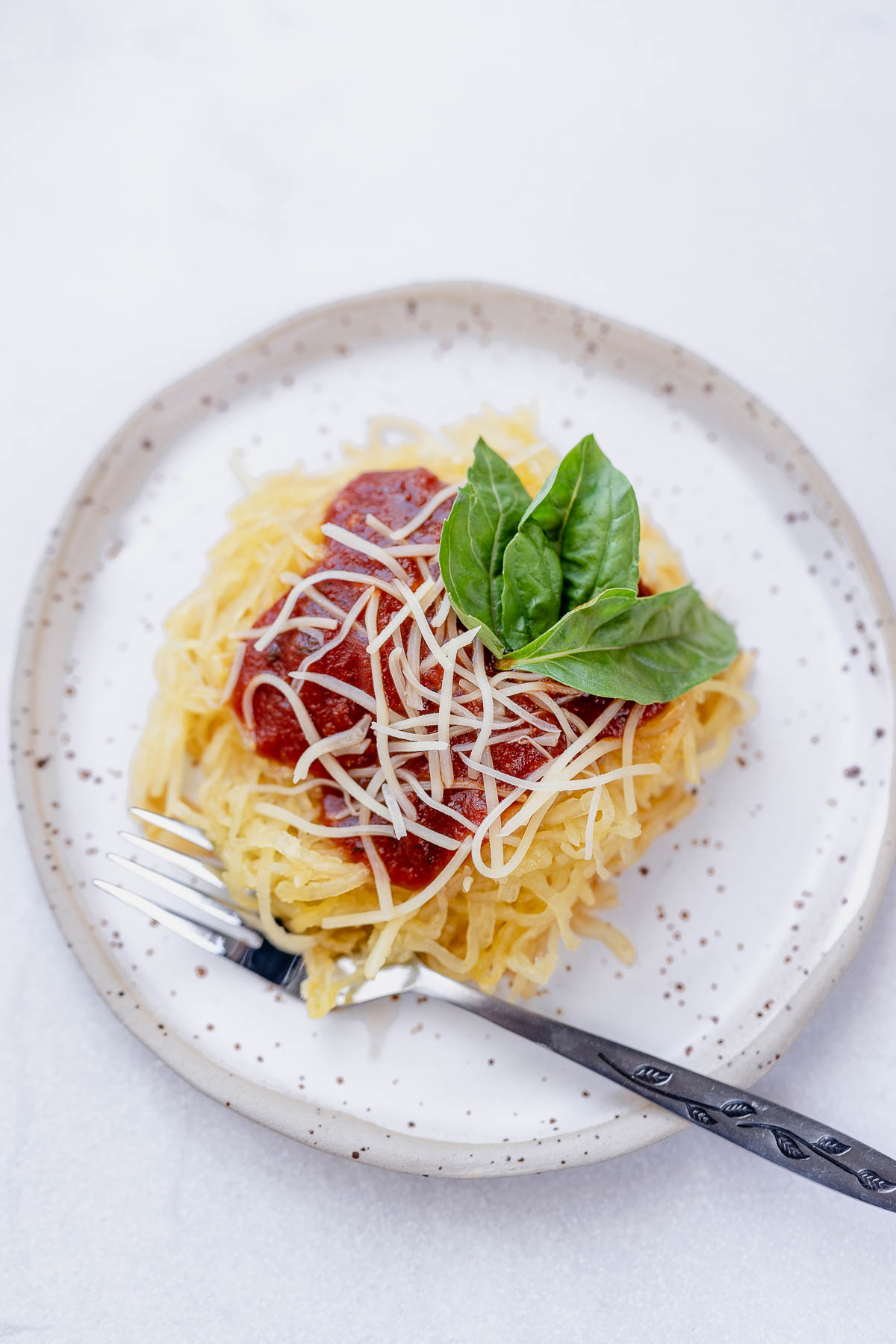 Overhead shot of Spaghetti squash on a stoneware plate with marinara, parmesan and fresh basil on top, and a fork.