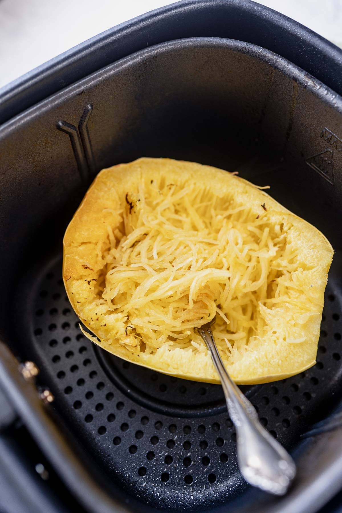 Fork pulling the flesh from half a cooked spaghetti squash in a black air fryer basket.