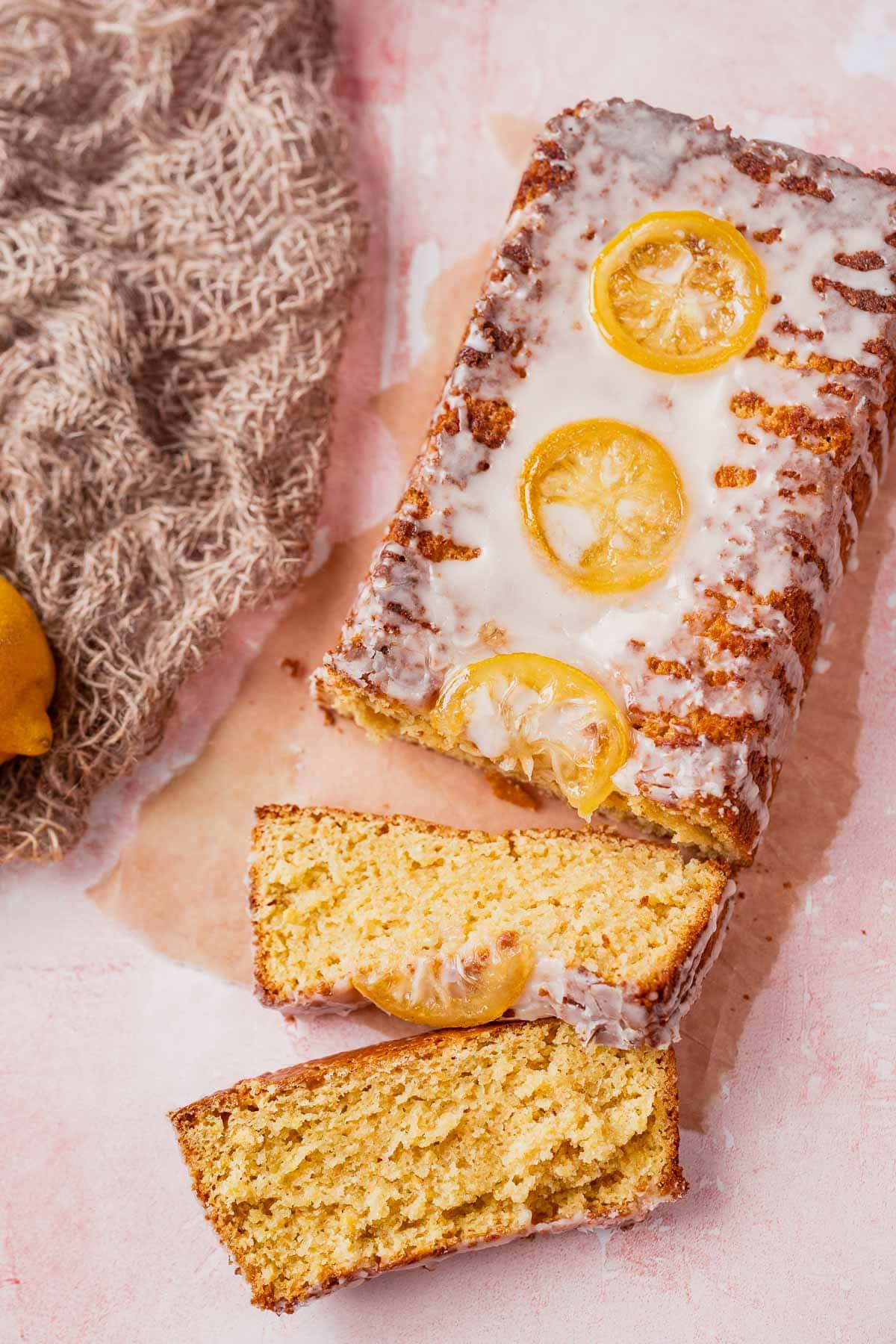 A sliced cake topped with white icing and lemon slices on a light pink table.