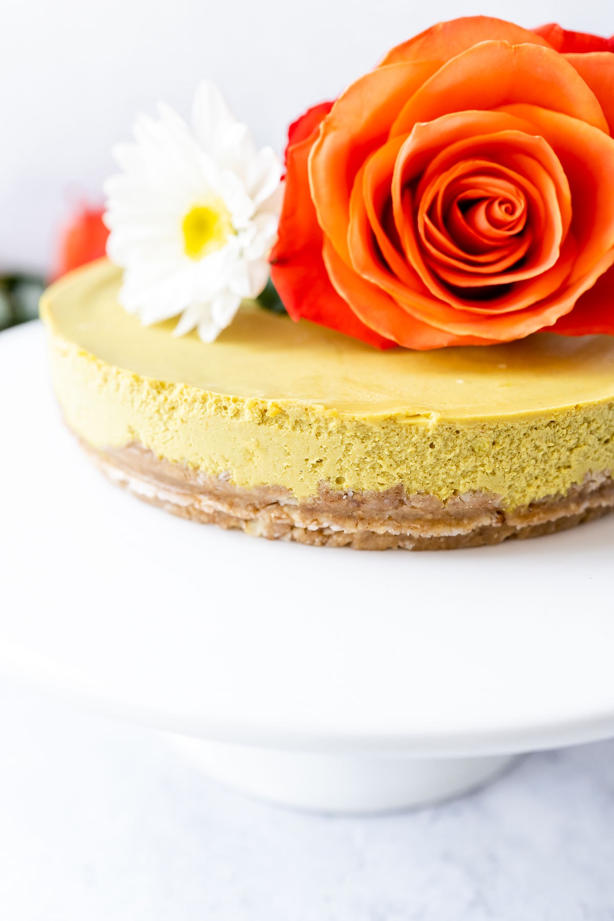 Close shot of a yellow vegan cheesecake topped with fresh flowers resting on a white cake stand.