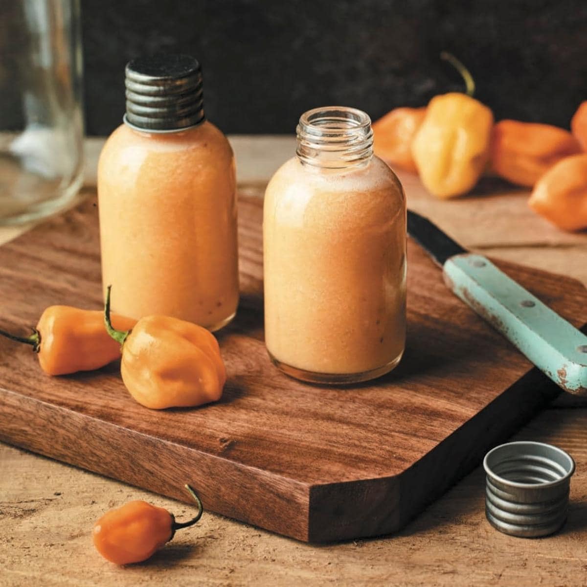 Clear glass bottles filled with orange red hot sauce resting on a wooden cutting board.