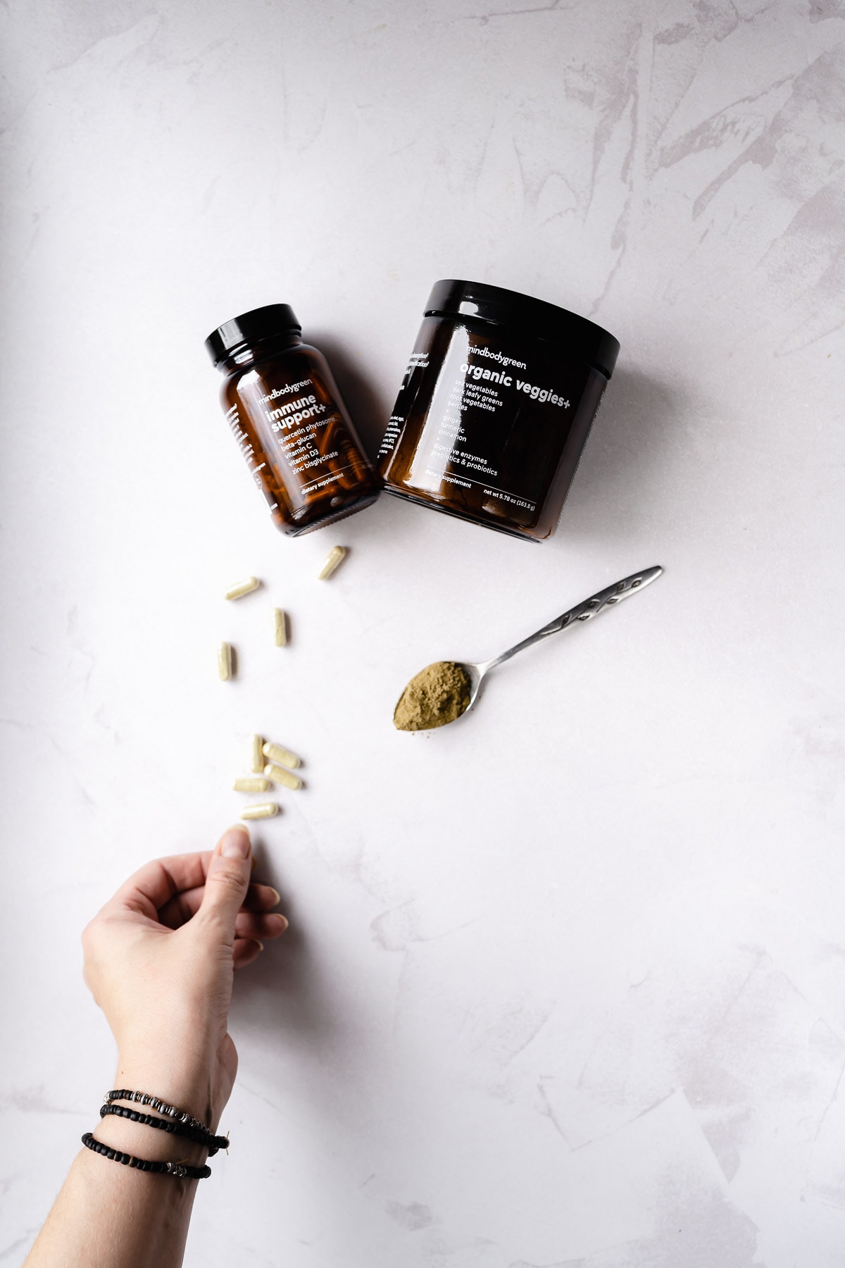 A hand reaches for a tan supplement capsule resting next to spoon of veggie powder.